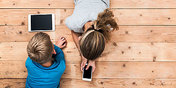 Zwei Kinder liegen auf dem Boden und beschäftigen sich mit ihrem Smartphone und Tablet.