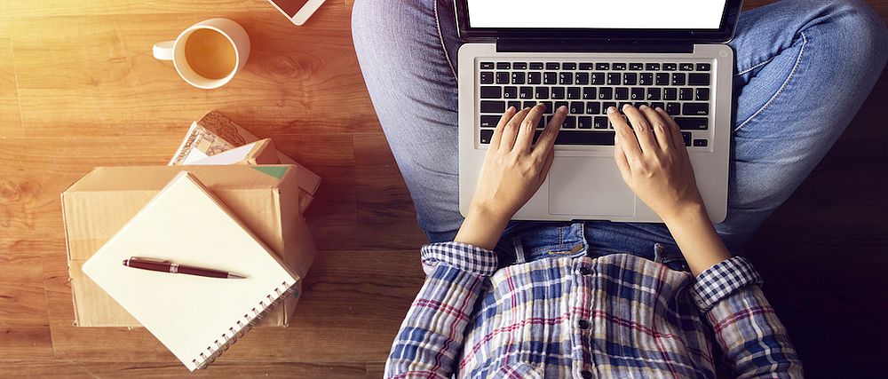 Eine Person sitzt auf dem Fußboden und tippt in ihr Notebook auf ihrem Schoß.