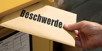 Eine Person wirft einen Briefumschlag mit der Aufschrift "Beschwerde" in einen Postbriefkasten.