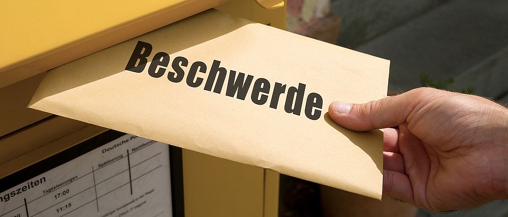 Eine Person wirft einen Briefumschlag mit der Aufschrift "Beschwerde" in einen Postbriefkasten.