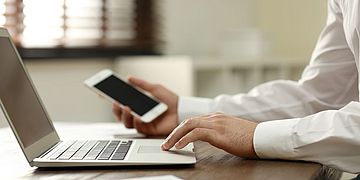 Eine Person hält in der einen Hand ein Smartphone und berührt mit der anderen Hand das Touchpad eines aufgeklappten Notebooks.