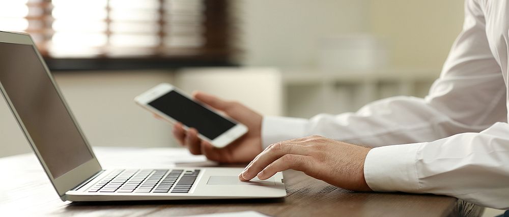 Eine Person hält in der einen Hand ein Smartphone und berührt mit der anderen Hand das Touchpad eines aufgeklappten Notebooks.