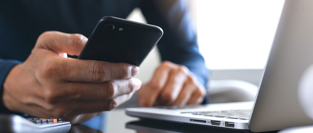 Eine Person blickt vor geöffnetem Notebook in ihr Smartphone.