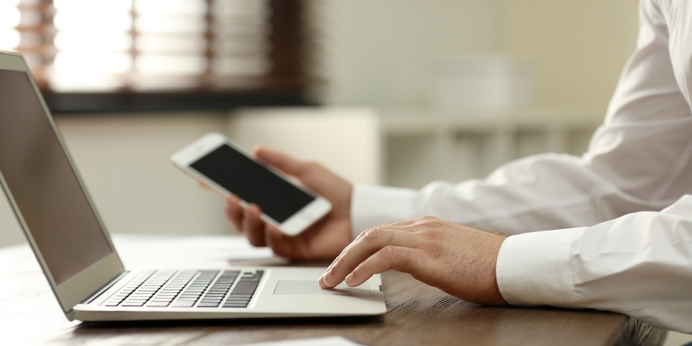 Eine Person hält in der einen Hand ein Smartphone und berührt mit der anderen Hand das Touchpad eines aufgeklappten Notebooks.